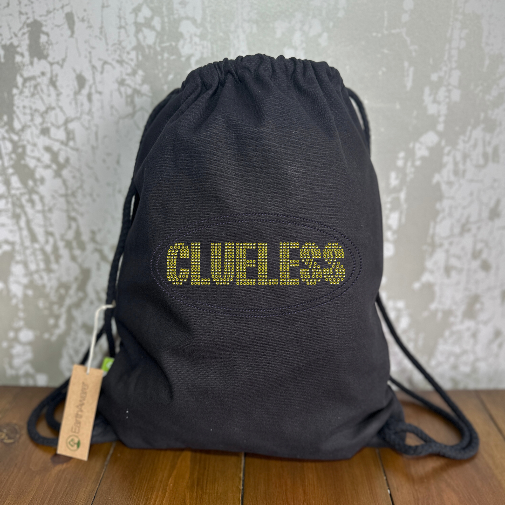 Thick cotton drawstring bag with bright yellow clueless writing in a block font, around the lettering is a black oval outline. The design is made up of sparkling rhinestones.
Design is placed on the front of the bag