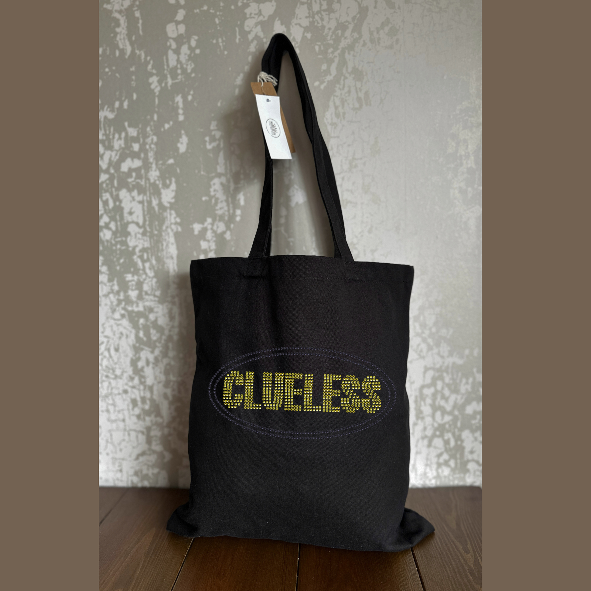 Black cotton tote bag with long handles, with bright yellow clueless writing in a block font, around the lettering is a black oval outline. The design is made up of sparkling rhinestones.
Design is placed on the front of the bag