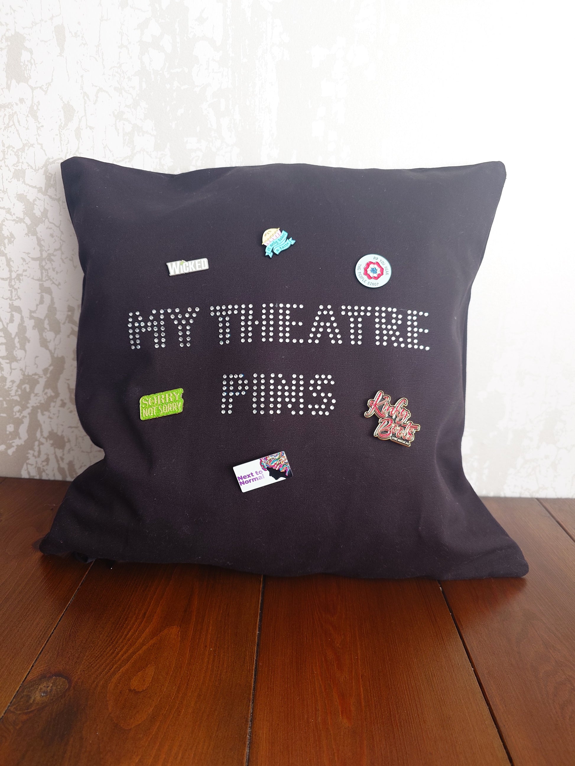Square black cotton cushion cover with silver rhinestone writing in the middle spelling out 'MY THEATRE PINS' the cushion has lots of theatre themed pins on it to show how it can be used to display pins