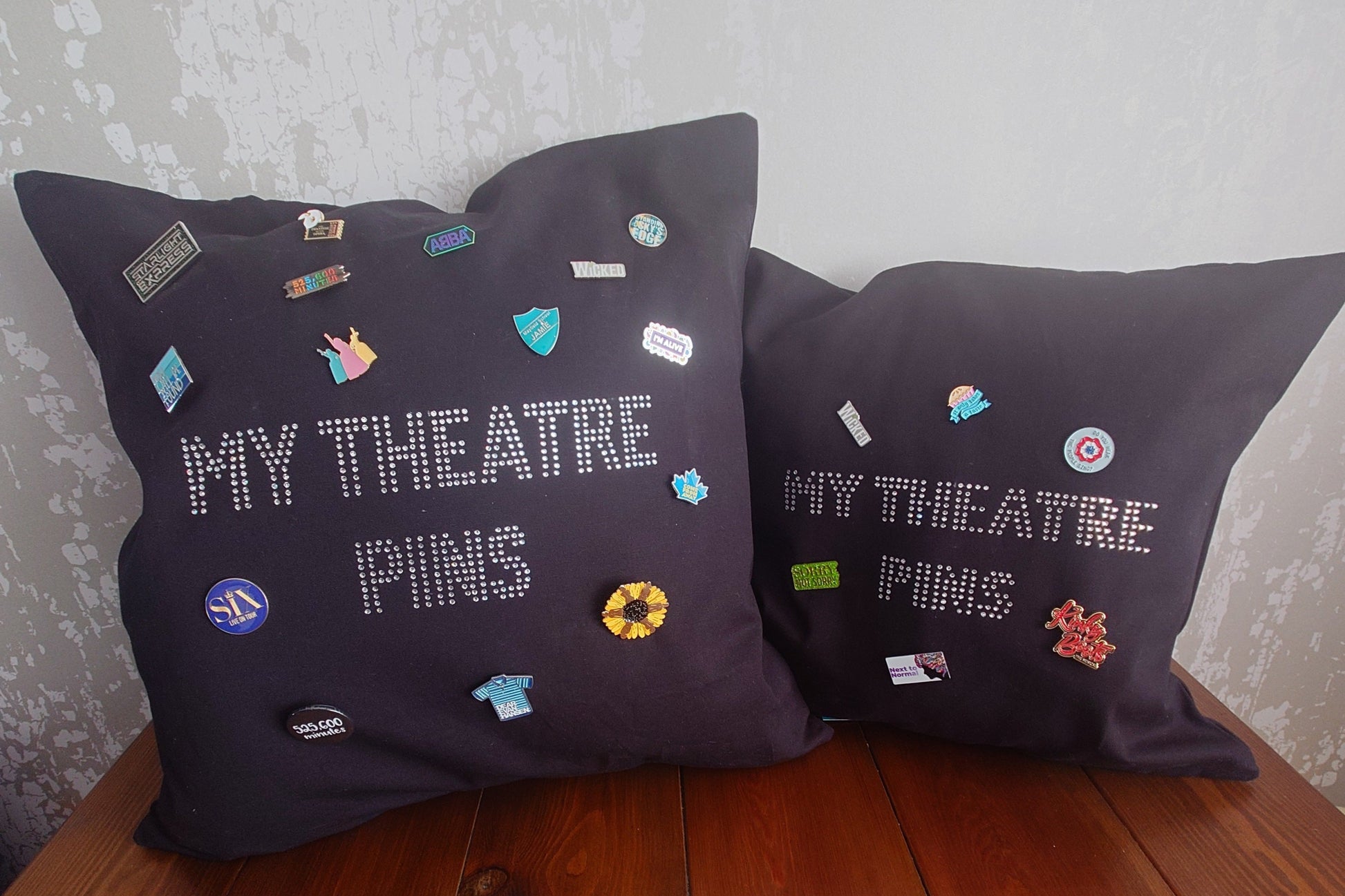 Two Square black cotton cushion cover with silver rhinestone writing in the middle spelling out 'MY THEATRE PINS' the cushion has lots of theatre themed pins on it to show how it can be used to display pins