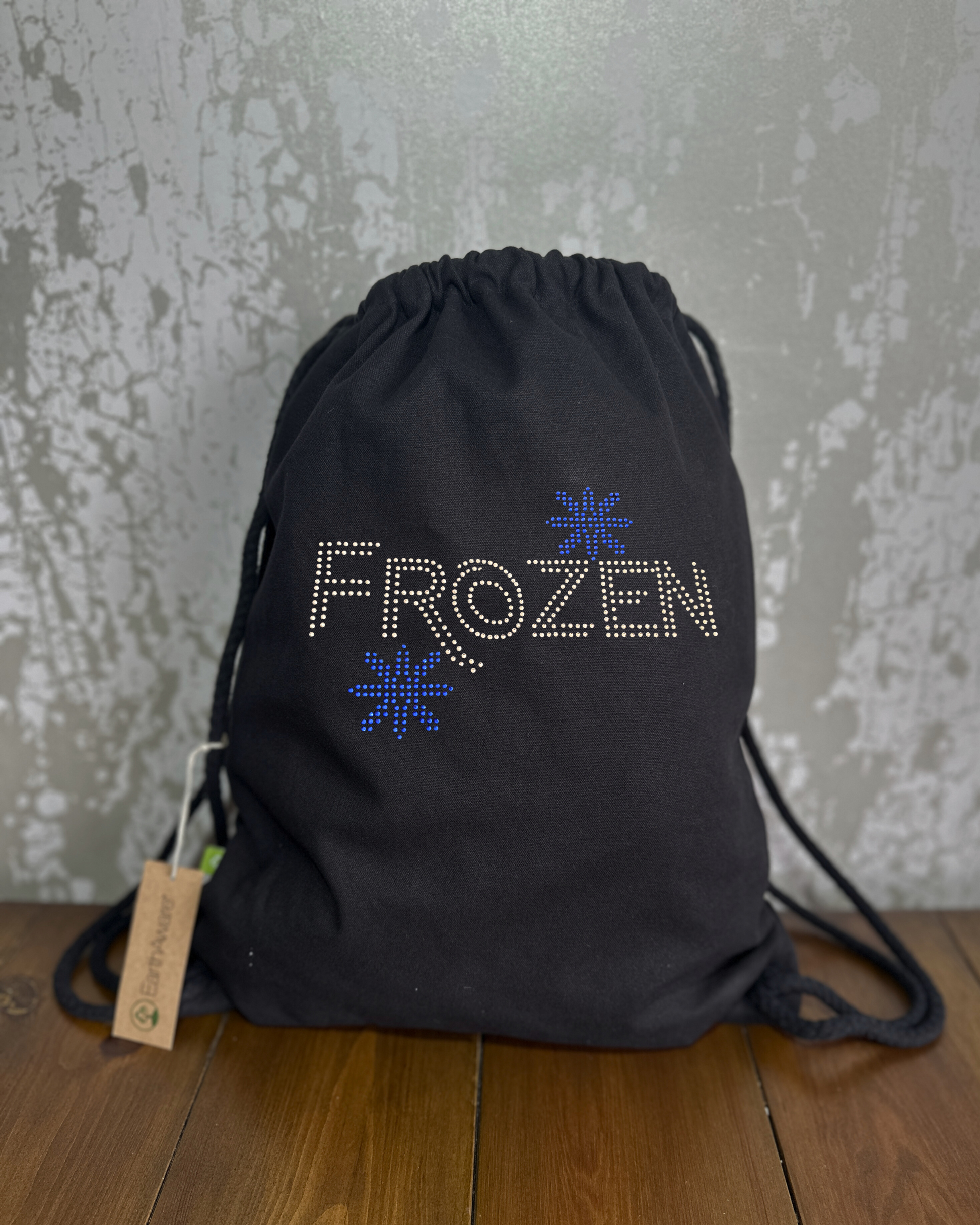 BLACK COTTON DRAWSTRING BAG MADE WITH THICK DURABLE MATERIAL. THE BAG HAS A CORD DRAWSTRING THAT ACT AS SHOULDER STRAPS. ON THE FRONT OF THE BAG IS  FROZEN LETTERING WITH TWO BLUE SNOWFLAKES SET ABOVE THE Z AND BELOW R. THE DESIGN IS MADE UP OF HOT FIX RHINESTONES THAT HEAT PRESSED AND CATCH THE LIGHT BEAUTIFULLY