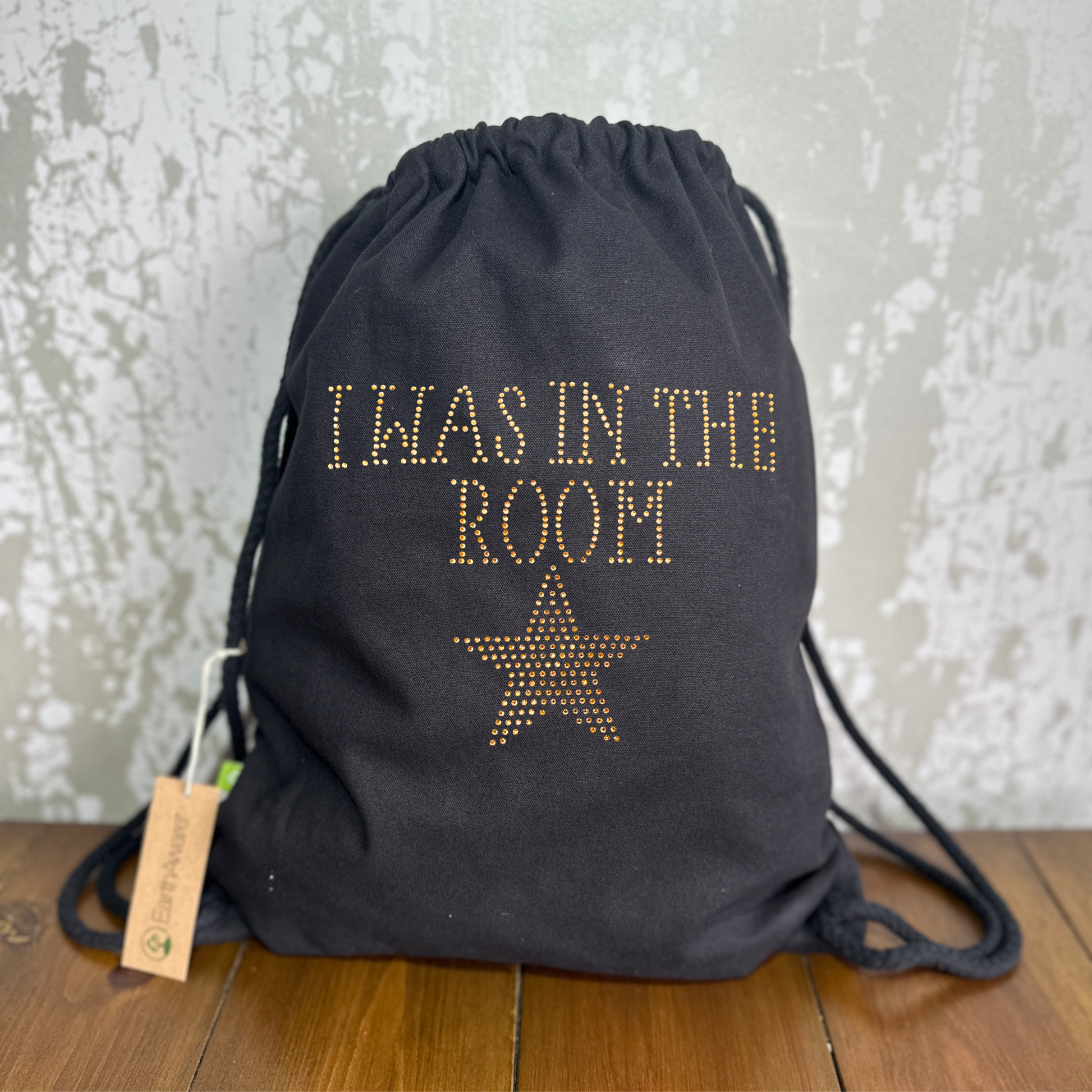 BLACK COTTON DRAWSTRING BAG MADE WITH THICK DURABLE MATERIAL. THE BAG HAS A CORD DRAWSTRING THAT ACT AS SHOULDER STRAPS. ON THE FRONT OF THE BAG IS  GOLD LETTERING SPELLING OUT 'I WAS IN THE ROOM" UNDERNEATH THIS IS GOLD STAR. THE DESIGN IS MADE UP OF HOT FIX RHINESTONES THAT HEAT PRESSED AND CATCH THE LIGHT BEAUTIFULLY