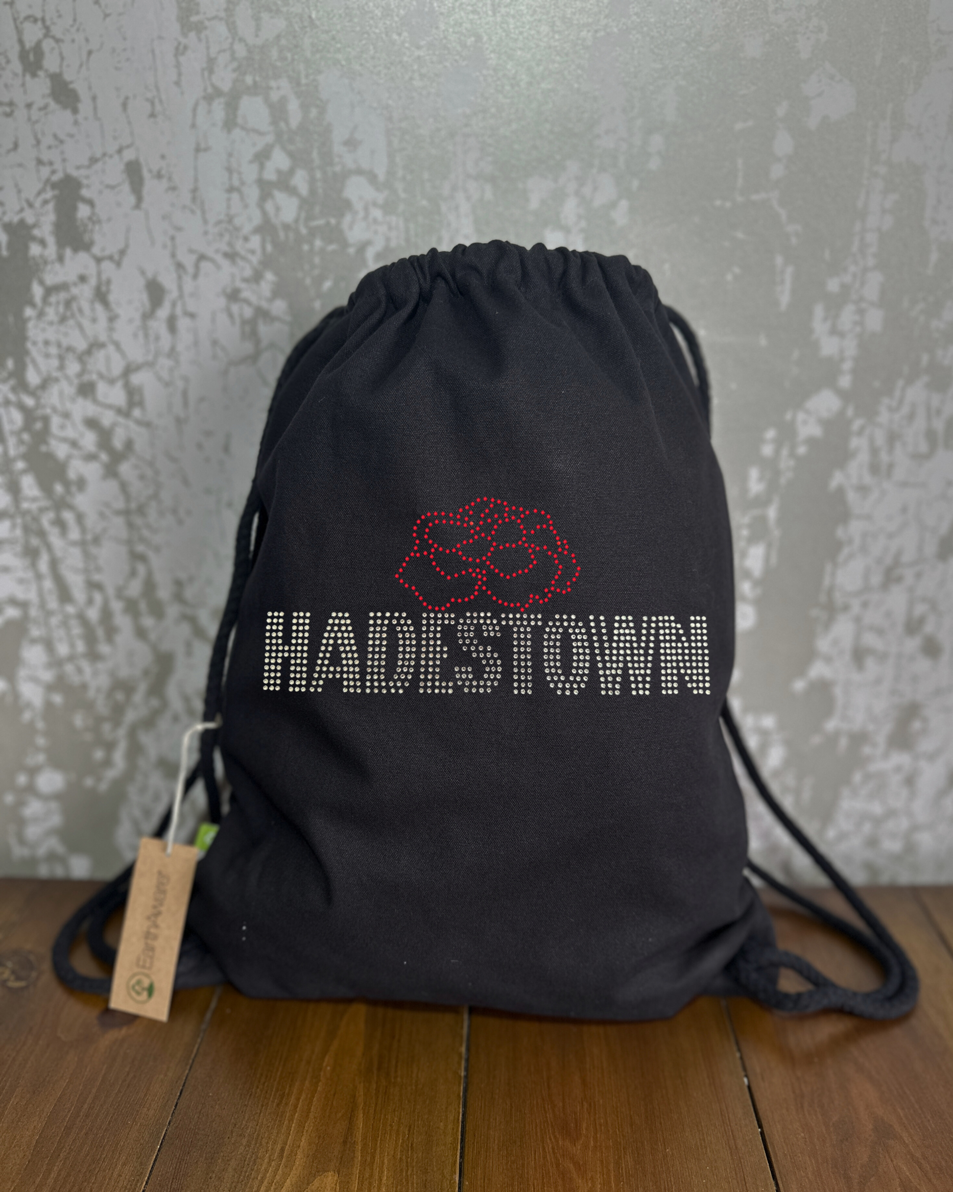 BLACK COTTON DRAWSTRING BAG MADE WITH THICK DURABLE MATERIAL. THE BAG HAS A CORD DRAWSTRING THAT ACT AS SHOULDER STRAPS. ON TH FRONT OF THE BAG IS A HADESTOWN DESIGN,  WITH SILVER SPARKLING HADESTOWN LETTERING AND THE RED SPARKLING OUTLINE OF A CARNATION. THE DESIGN IS MADE UP OF HOT FIX RHINESTONES THAT CATCH THE LIGHT BEAUTIFULLY
