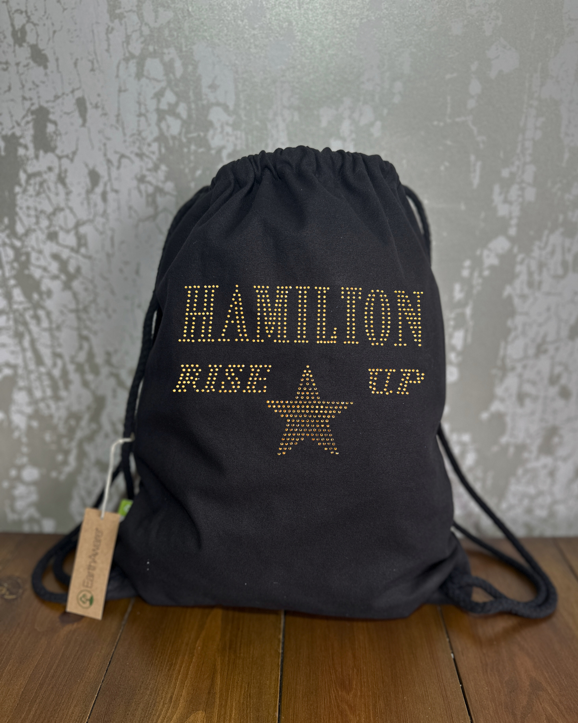 BLACK COTTON DRAWSTRING BAG MADE WITH THICK DURABLE MATERIAL. THE BAG HAS A CORD DRAWSTRING THAT ACT AS SHOULDER STRAPS. ON THE FRONT OF THE BAG IS  GOLD LETTERING SPELLING OUT 'HAMILTON RISE UP" UNDERNEATH THIS IS GOLD STAR. THE DESIGN IS MADE UP OF HOT FIX RHINESTONES THAT HEAT PRESSED AND CATCH THE LIGHT BEAUTIFULLY