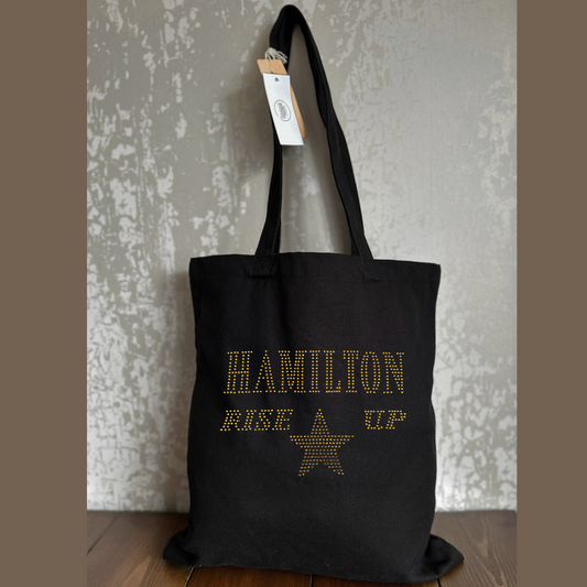 Black cotton tote bag, on the front is a gold sparkling "Hamilton Rose up" lettering with a gold star set just underneath.
design is made up of sparkling rhinestones that catch the light beautifully