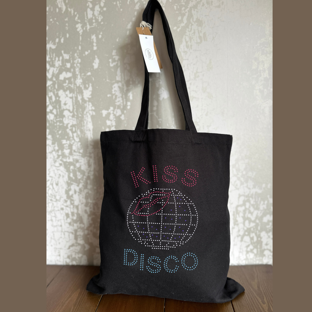 Black cotton tote bag with harry styles themed design on the front. 
KISS spelt out in pink rhinestones this curves round the top of a silver disco ball with a pink lip design and purple stones embedded it to the ball design. the word Disco is then curved underneath in aquamarine blue.