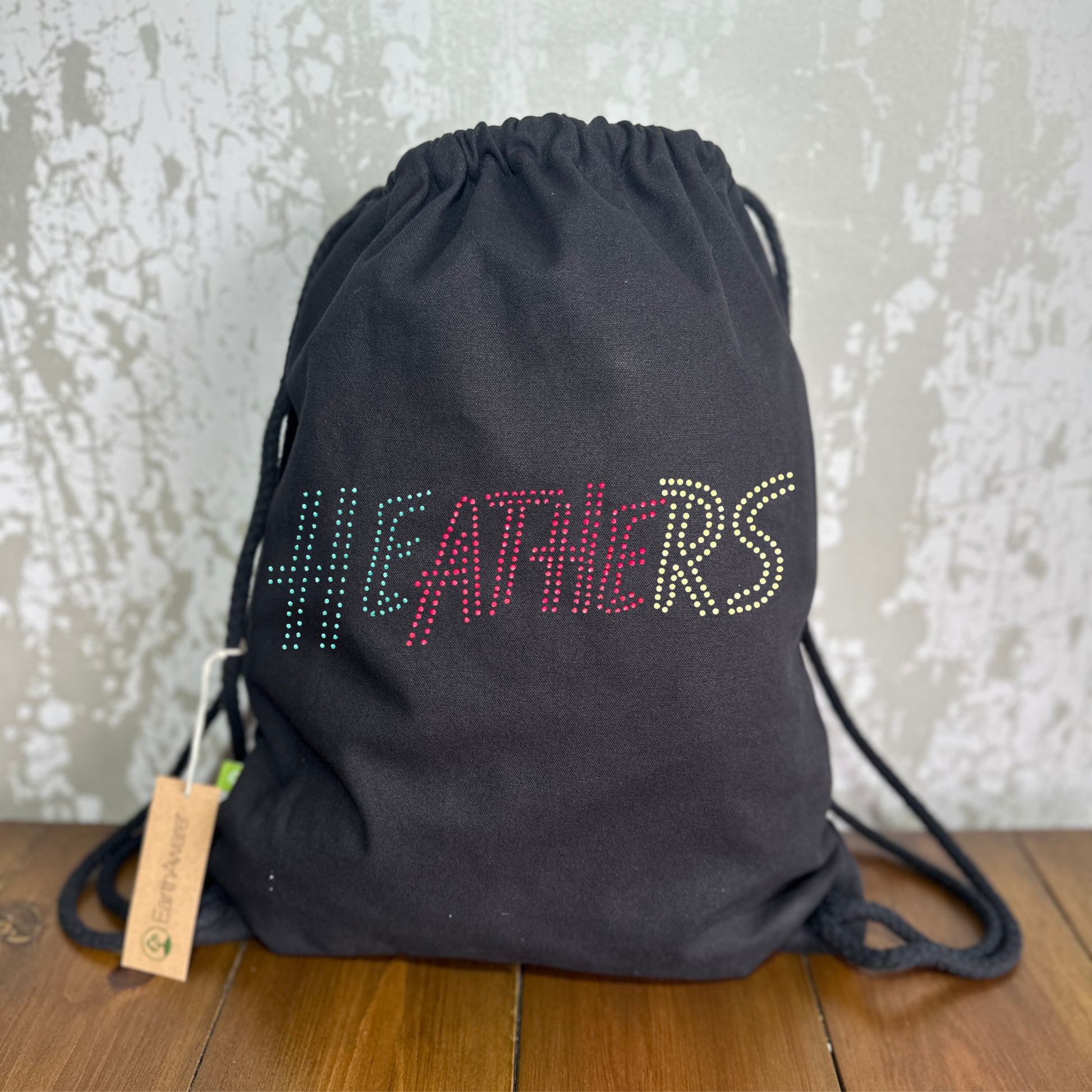 BLACK COTTON DRAWSTRING BAG MADE WITH THICK DURABLE MATERIAL. THE BAG HAS A CORD DRAWSTRING THAT ACT AS SHOULDER STRAPS. ON TH FORNT OF THE BAG IS A HEATHERS INSPIRED DEISGN WITH LETTERING SPELLING OUT HEATHERS IN FIRST GREEN, THEN RED, THEN YELLOW