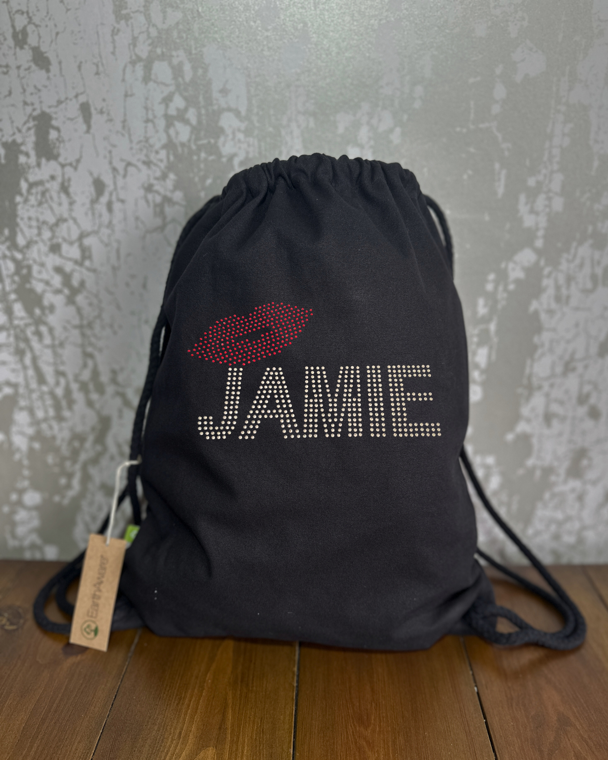 EVERYBODY'S TALKING ABOUT JAMIE, BLACK COTTON DRAWSTRING BAG MADE WITH THICK DURABLE MATERIAL. THE BAG HAS A CORD DRAWSTRING THAT ACT AS SHOULDER STRAPS. ON THE FRONT OF THE BAG IS  SILVER LETTERING SPELLING OUT 'THE NAME JAMIE, SET JUST ABOVE THE J IS SET OF RED LIPS. THE DESIGN IS MADE UP OF HOT FIX RHINESTONES THAT HEAT PRESSED AND CATCH THE LIGHT BEAUTIFULLYS 