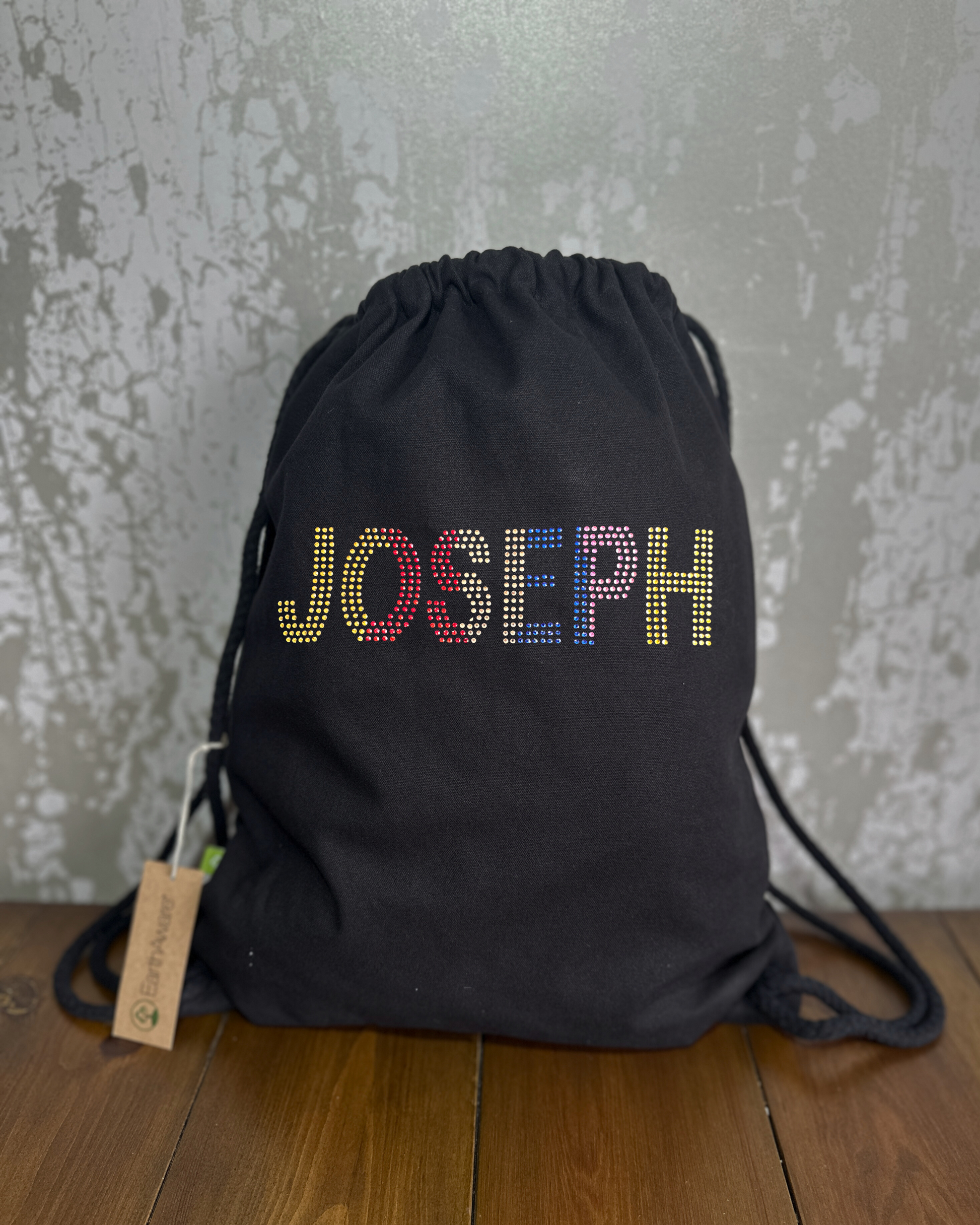 BLACK COTTON DRAWSTRING BAG MADE WITH THICK DURABLE MATERIAL. THE BAG HAS A CORD DRAWSTRING THAT ACT AS SHOULDER STRAPS. ON THE FRONT OF THE BAG IS  IS THE WORD JOSEPH. THIS WORD GRADUALLY CHANGES FROM GOLD TO RED, CHAMPAGNE, BLUE, PURPLE AND BACK TO GOLD, THE DESIGN IS MADE UP OF HOT FIX RHINESTONES THAT HEAT PRESSED AND CATCH THE LIGHT BEAUTIFULLY