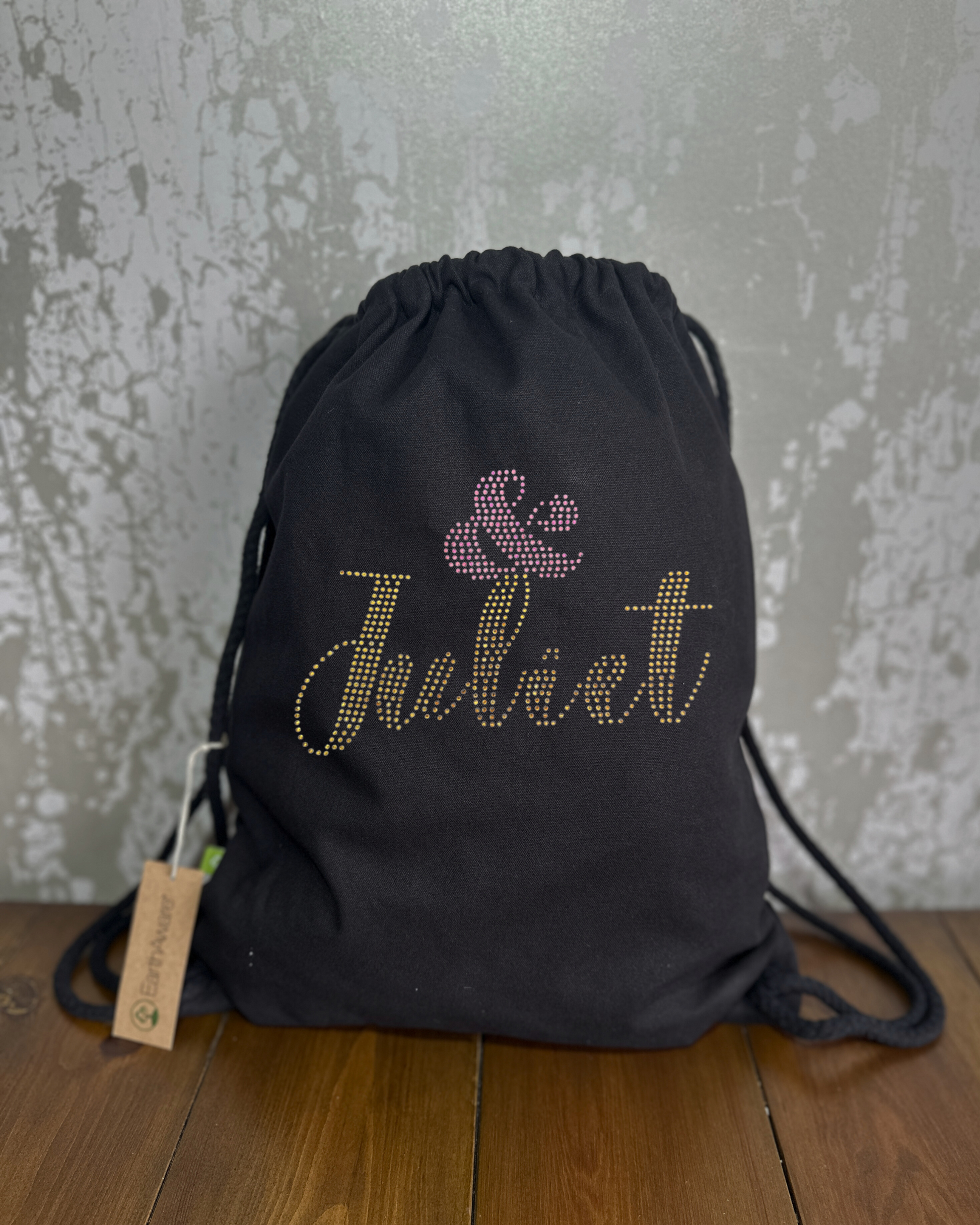 BLACK COTTON DRAWSTRING BAG MADE WITH THICK DURABLE MATERIAL. THE BAG HAS A CORD DRAWSTRING THAT ACT AS SHOULDER STRAPS. ON THE FRONT OF THE BAG IS  AN &JULIET DESIGN IN GOLD AND PINK, THE NAME JULIET IS IN GOLD AND THERE IS PINK & SYMBOL SET JUST ABOVE THE L OF JULIET. THE DESIGN IS MADE UP OF HOT FIX RHINESTONES THAT HEAT PRESSED AND CATCH THE LIGHT BEAUTIFULLY