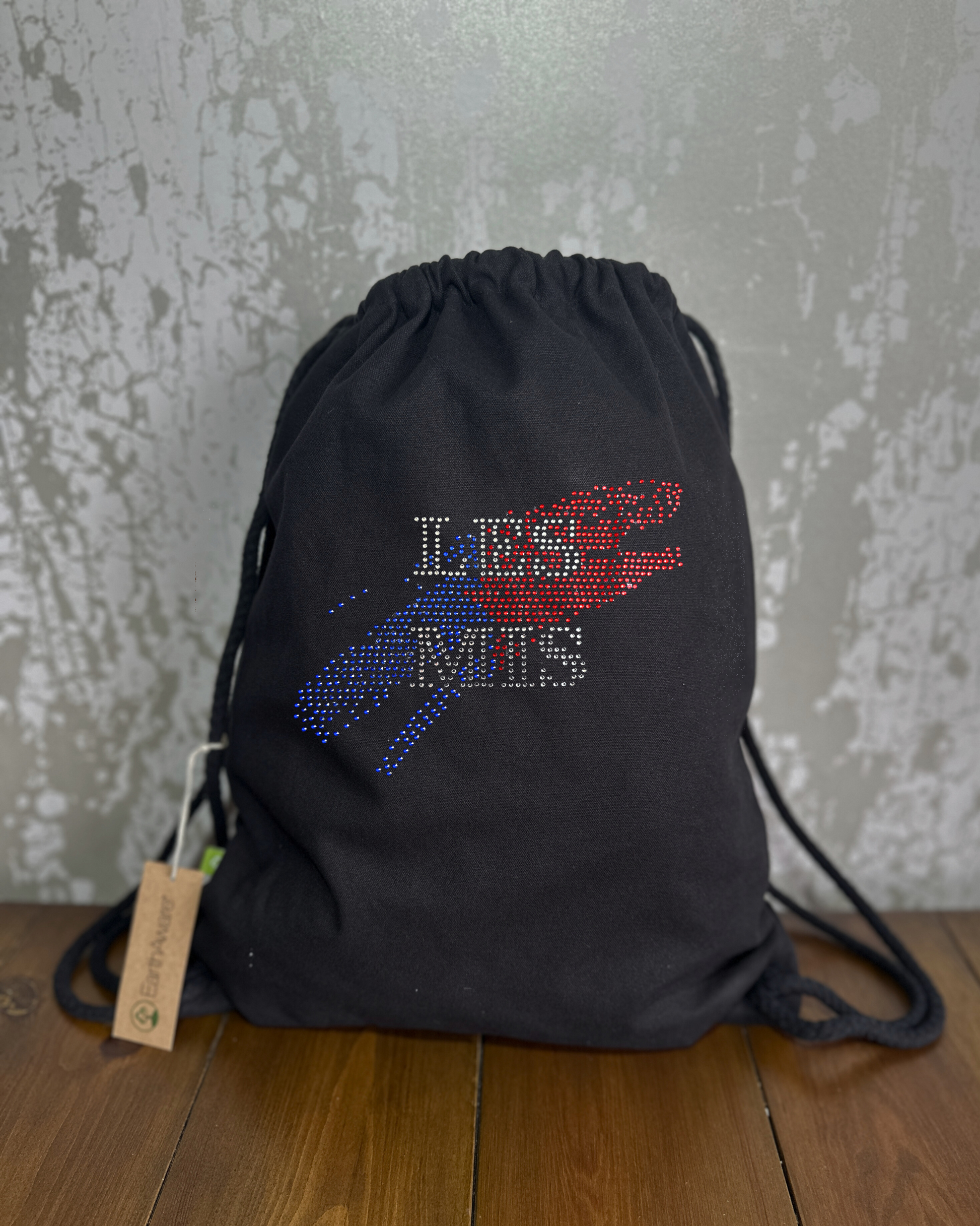 BLACK COTTON DRAWSTRING BAG MADE WITH THICK DURABLE MATERIAL. THE BAG HAS A CORD DRAWSTRING THAT ACT AS SHOULDER STRAPS. ON THE FRONT OF THE BAG IS  SILVER LES MIS WRITING SET INTO A BLUE AND RED SPLASH OF COLOUR. THE DESIGN IS MADE UP OF HOT FIX RHINESTONES THAT HEAT PRESSED AND CATCH THE LIGHT BEAUTIFULLY