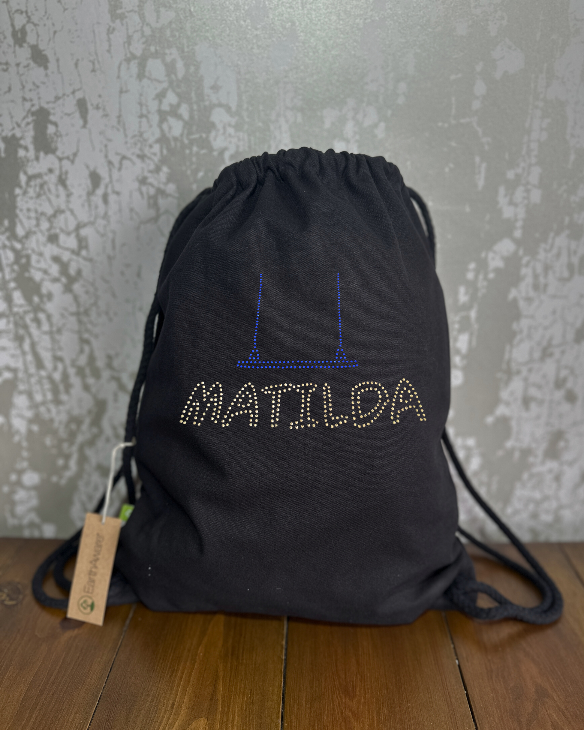 MATILDA BLACK COTTON DRAWSTRING BAG MADE WITH THICK DURABLE MATERIAL. BLUE SWING DETAIL WITH SILVER MATILDA LETTERING, THE BAG HAS A CORD DRAWSTRING THAT ACT AS SHOULDER STARPS. 
THE DESIGN IS MADE UP OF HOT FIX RHINESTONES THAT HEAT PRESSED AND CATCH THE LIGHT BEAUTIFULLY