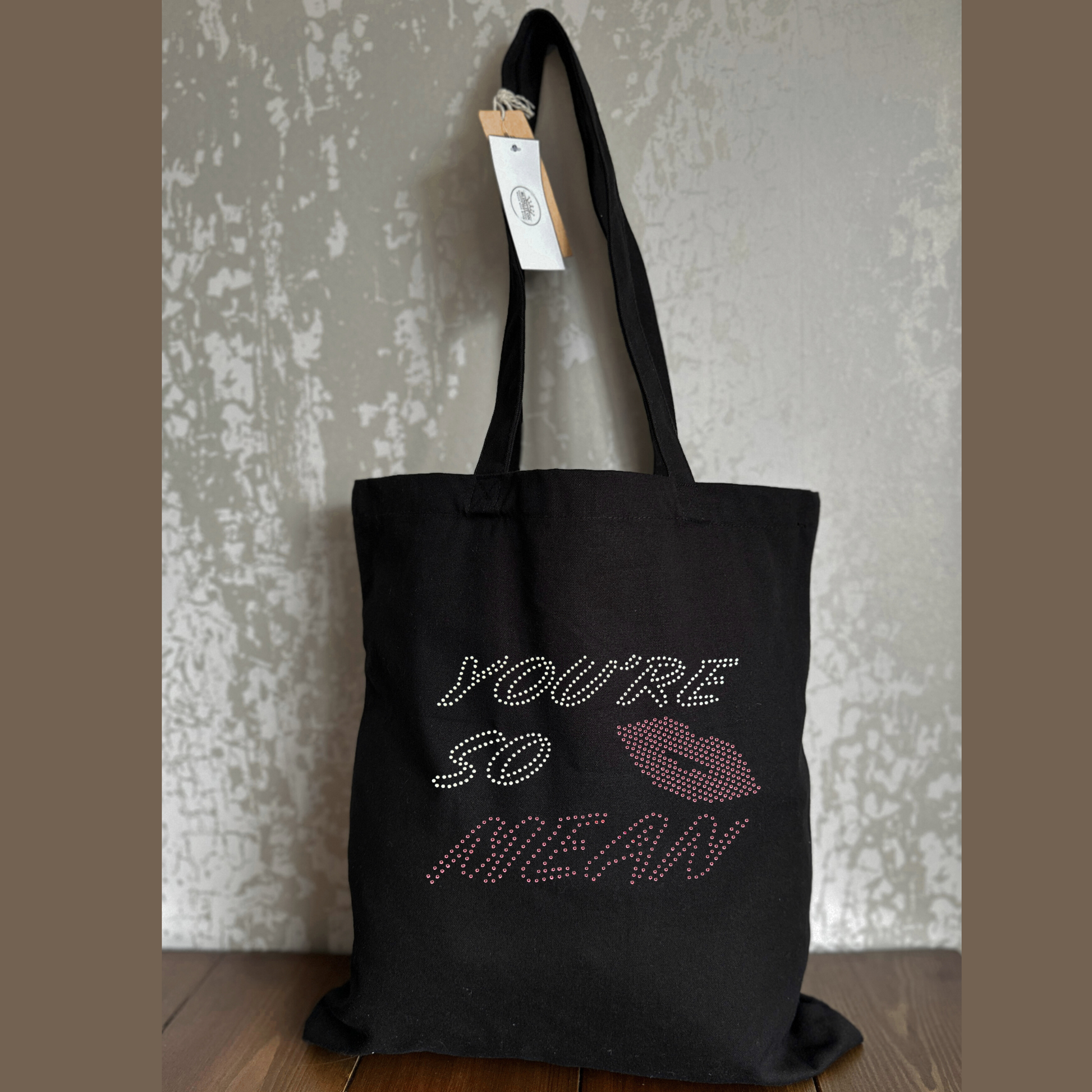Mean girls, Black cotton heavy tote bag with long handles, on the front of the t shirt, is silver and pink sparkly writing spelling out 'You're so mean" the last word mean is in pink, set in the middle of the lettering is a set of pink lips.