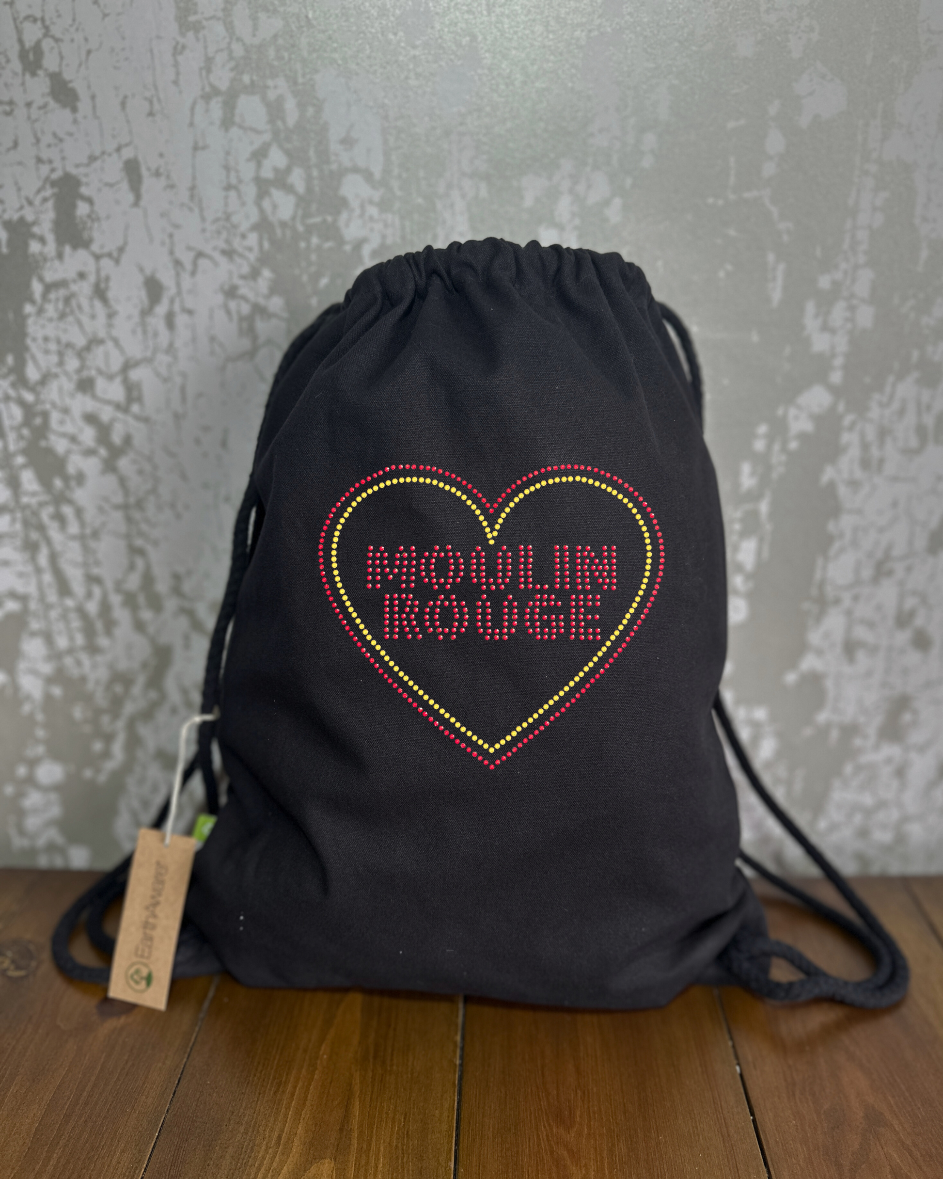 BLACK COTTON DRAWSTRING BAG MADE WITH THICK DURABLE MATERIAL. THE BAG HAS A CORD DRAWSTRING THAT ACT AS SHOULDER STRAPS. ON TH FRONT OF THE BAG IS A MOULIN ROUGE DESIGN, THERE IS RED SPARKLING LETTERING WITH GOLD AND RED HEART AROUND THE WRITING. THE DESIGN IS MADE UP OF HOT FIX RHINESTONES THAT HEAT PRESSED AND CATCH THE LIGHT BEAUTIFULLY