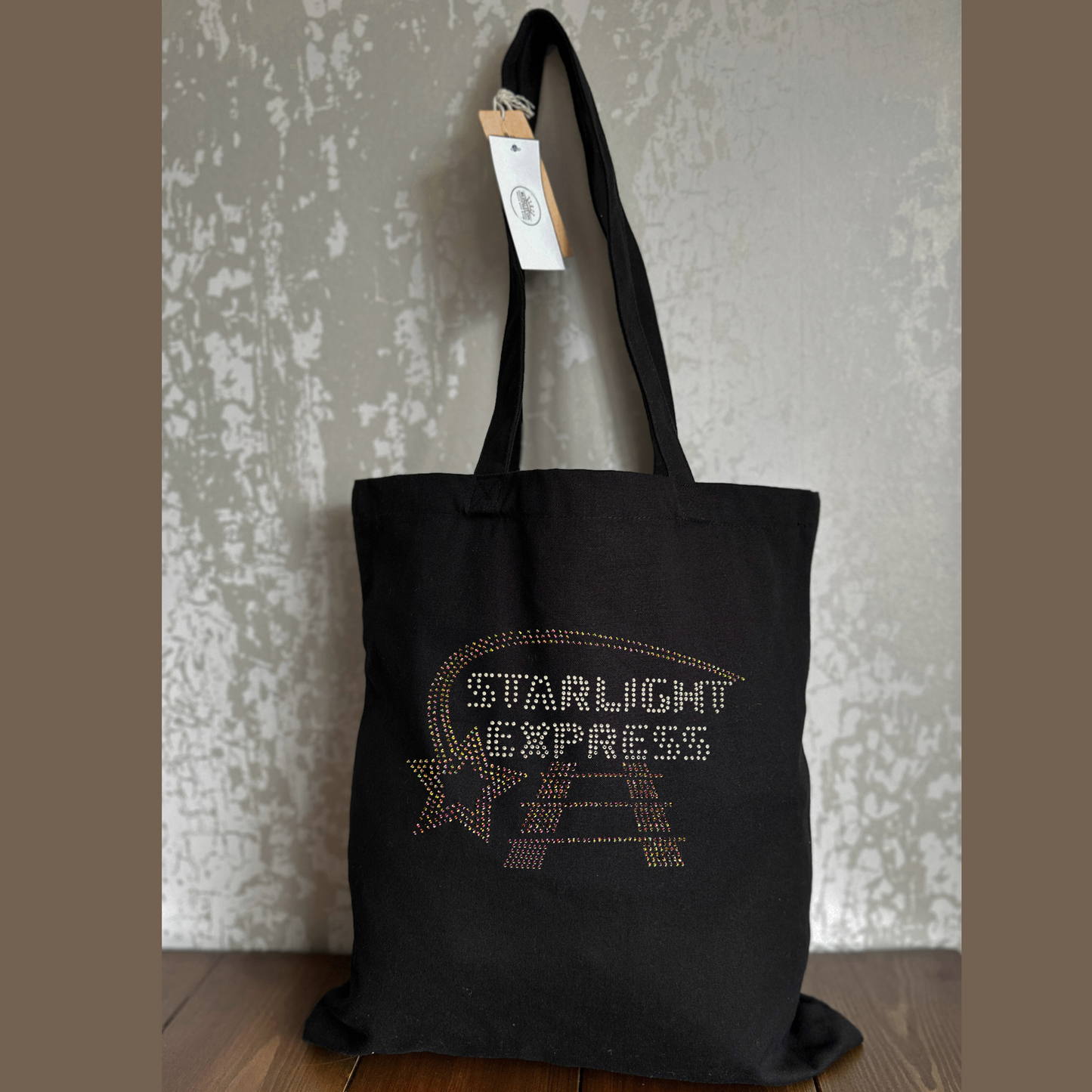 Black cotton tote bag, on the front there is  silver Starlight Express writing with rainbow rose gold starlight trail over the top of the writing and train track underneath.
The design is made up of sparkling hot fix rhinestones that catch the light and sparkle.