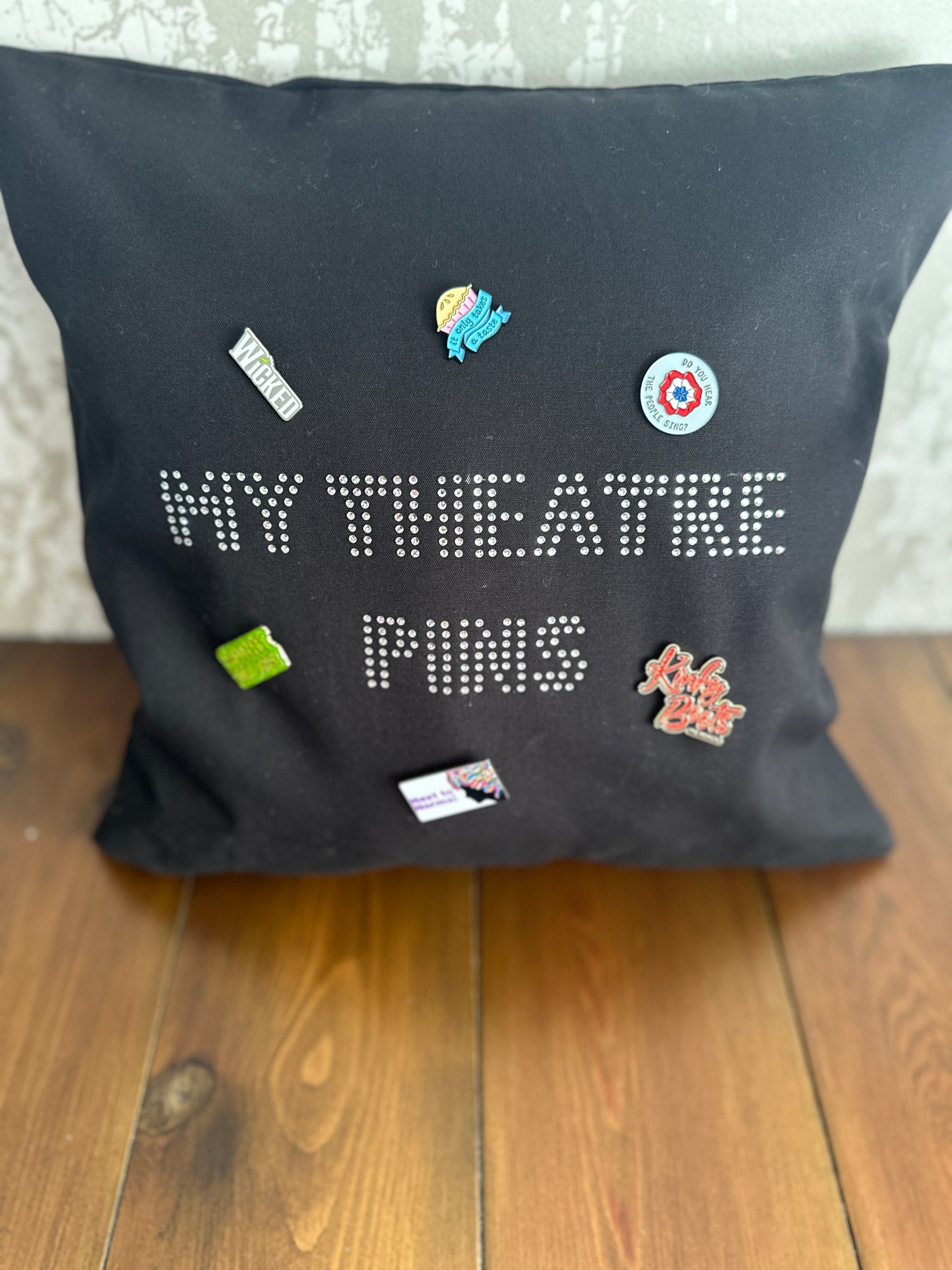 Square black cotton cushion cover with silver rhinestone writing in the middle spelling out 'MY THEATRE PINS' the cushion has lots of theatre themed pins on it to show how it can be used to display pins