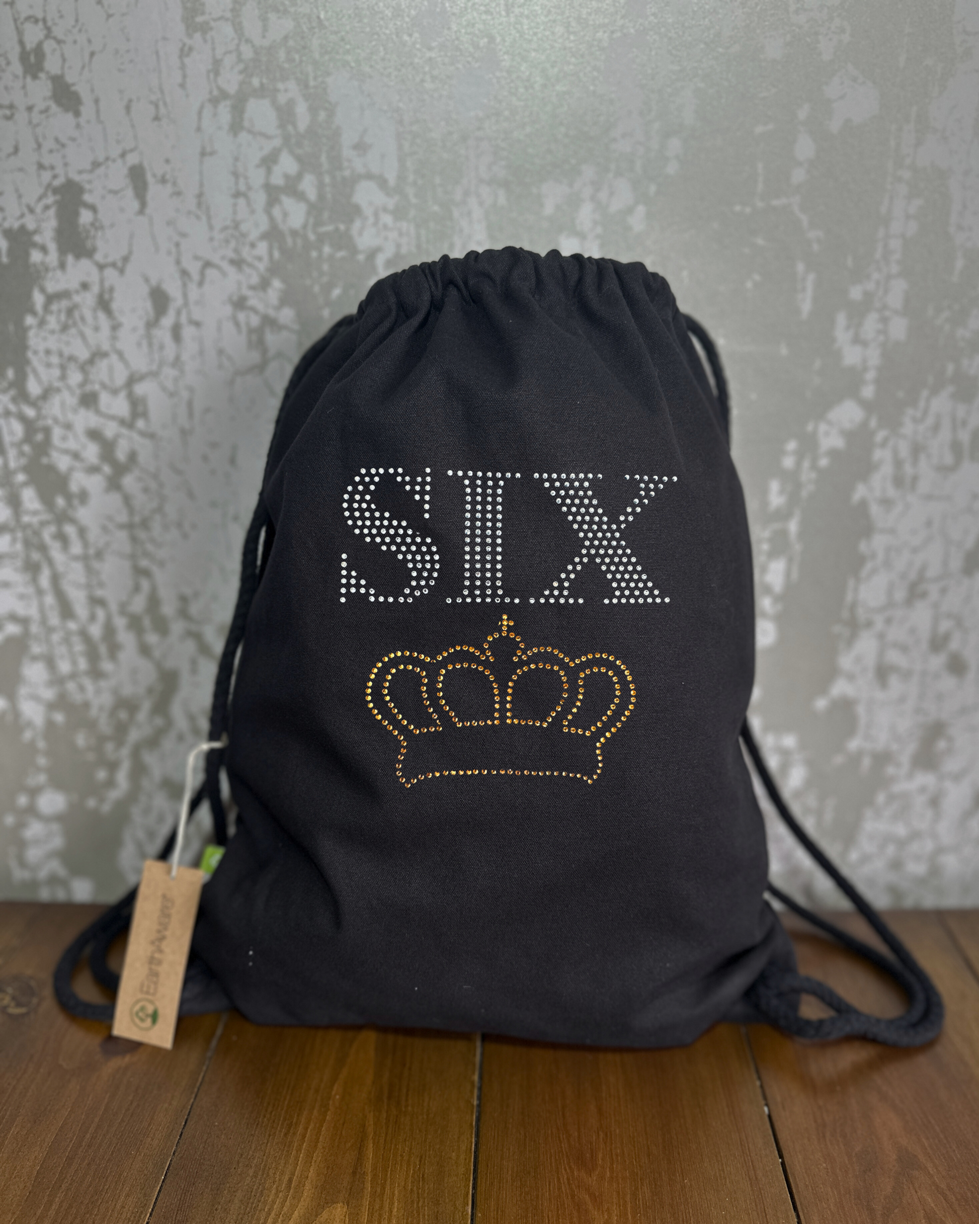 BLACK COTTON DRAWSTRING BAG MADE WITH THICK DURABLE MATERIAL. THE BAG HAS A CORD DRAWSTRING THAT ACT AS SHOULDER STRAPS. ON THE FRONT OF THE BAG IS SIX LETTERING AND GOLDEN CROWN UNDERNEATH. THE DESIGN IS MADE UP OF HOT FIX RHINESTONES THAT HEAT PRESSED AND CATCH THE LIGHT BEAUTIFULLY
