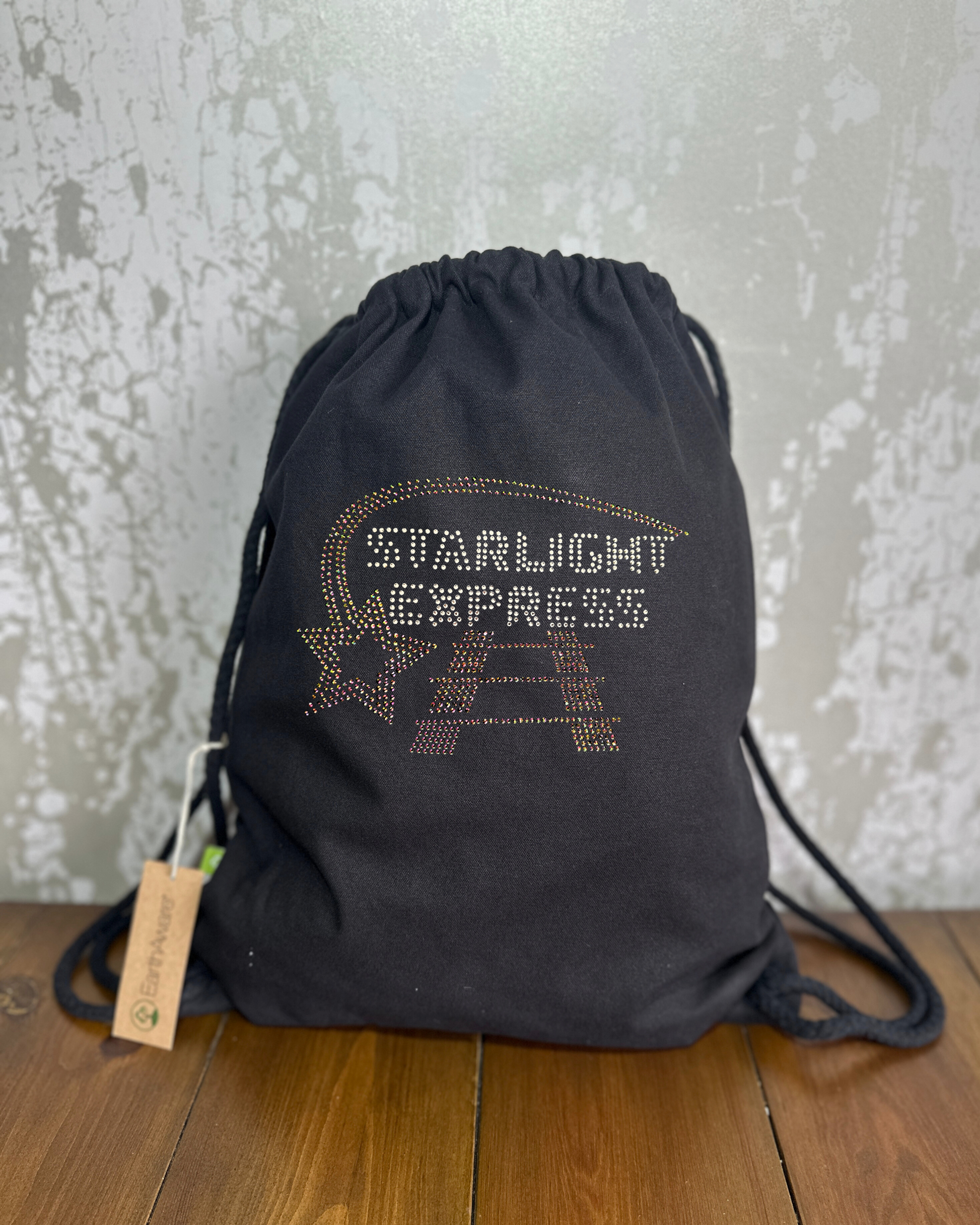 BLACK COTTON DRAWSTRING BAG MADE WITH THICK DURABLE MATERIAL. THE BAG HAS A CORD DRAWSTRING THAT ACT AS SHOULDER STRAPS. ON THE FRONT OF THE BAG IS SILVER LETTERING SPELLING OUT STARLIGHT EXPRESS, ACROSS THE TOP OF THIS LETTERING IS A RAINBOW ROSE GOLD STAR TRAIL AND UNDERNEATH A TRAIN TRACK. THE DESIGN IS MADE UP OF HOT FIX RHINESTONES THAT HEAT PRESSED AND CATCH THE LIGHT BEAUTIFULLY